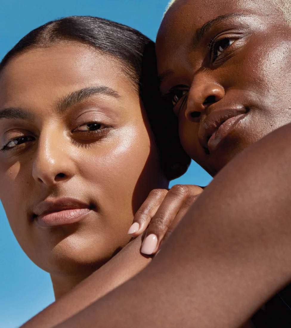 A close-up portrait of two individuals with diverse skin tones. One has warm medium-toned skin looking directly forward, while the other with dark skin and short blonde hair looks upwards, both showcasing healthy, glowing complexions against a blue sky.